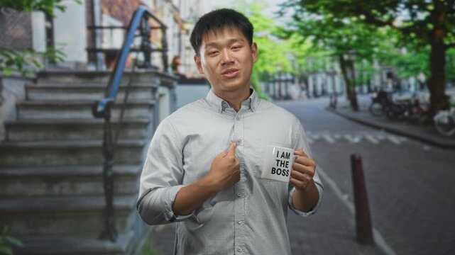 Man points finger to mug reading i am the boss while holding cup near chest, standing by stone steps and parked bicycles on a tree lined street; playful confidence.