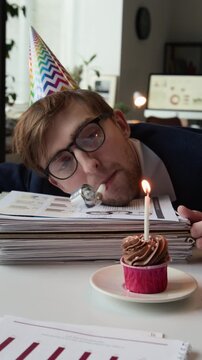 Vertical view of exhausted Caucasian gen Z office worker with multicoloured hat blowing into party blower resting head on pile of documents next to birthday cupcake with burning candle