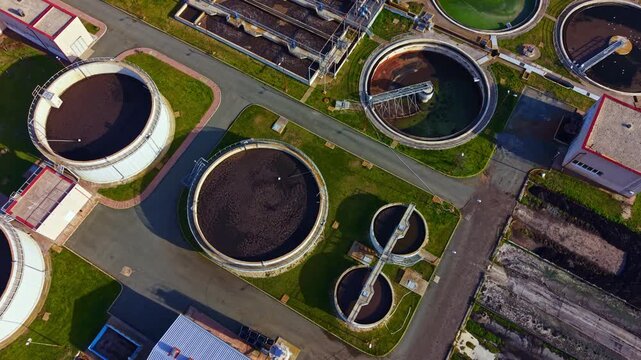 A view from above shows a wastewater treatment facility with multiple round tanks and processing areas. The facility is set in a clear environment with structures and pathways.