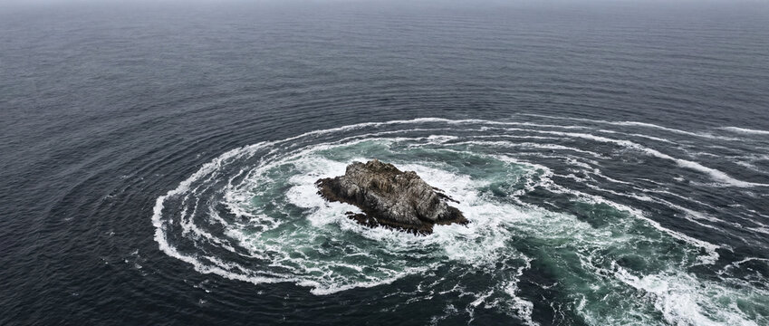 Aerial view of a lonely rocky sea stack encircled by swirling tidal currents and white ocean foam, set in dark open water under an overcast sky with dramatic natural patterns from above
