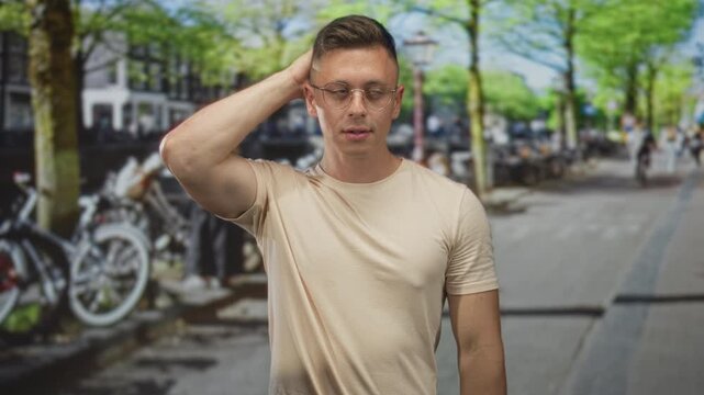 Man touching neck with hand behind head wearing beige tshirt on street by parked bicycles and trees; thoughtful introspection.