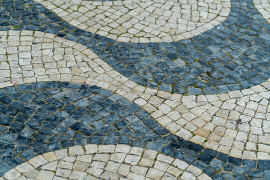 Intricate calcada patterns in Portugal showcase blend of light and dark stones arranged in wavy designs. Unique flooring Calcada enhances the charm of the surrounding area.