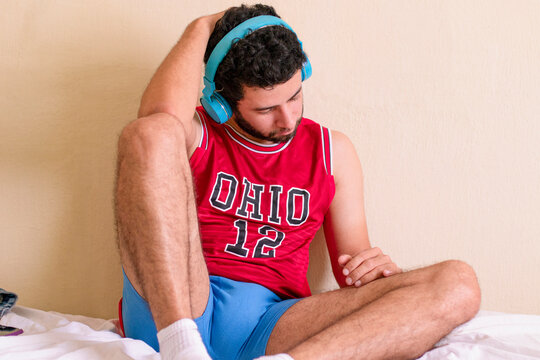 Man sitting on the bed relaxing listening to music, man sitting on the bed listening to music on headphones. Handsome bearded man with a fit body, red team shirt and blue underwear. Legs and arms