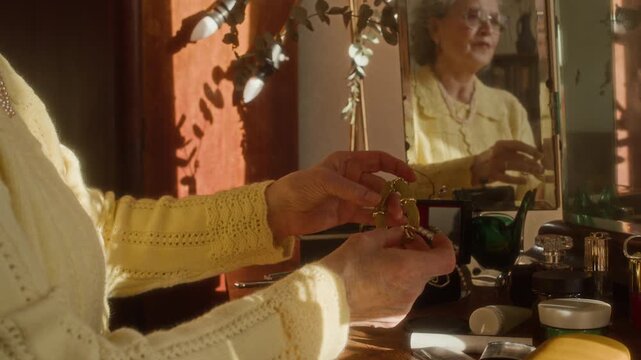 Side view of senior woman with white hair sitting at vanity table trying on vintage bracelet while getting ready in sunlit bedroom