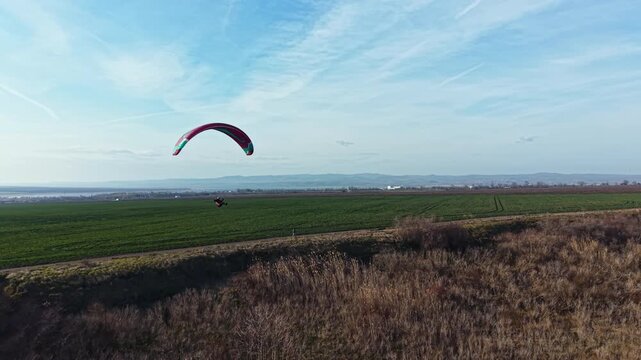 A paraglider soars through the sky above a green field with hills in the distance on a clear day. The scene shows open space and natural beauty.