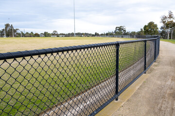 Obraz premium Chain link fence along sports field with grassy oval and walking path in suburban Melbourne Australia. Community sports infrastructure. Enclosed public recreational space for local activity
