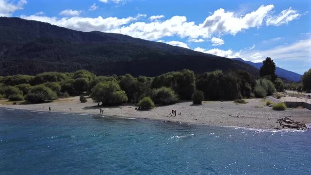 Peaceful shot of a pebble beach on the shore of Lago Puelo in Patagonia Chubut. Crystalline water, native vegetation, and the Andes mountains are visible on a sunny day.