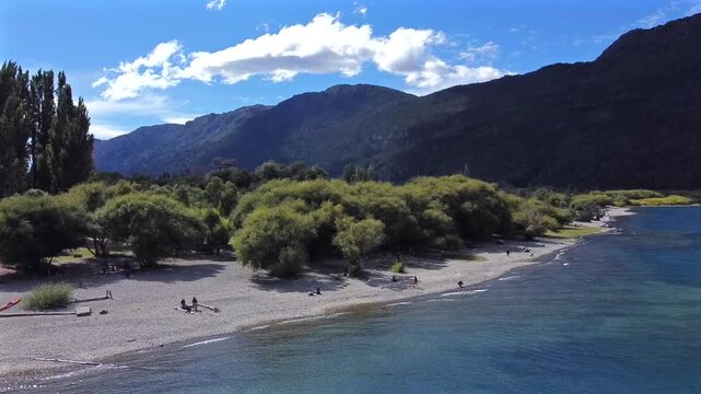 Aerial shot from the water towards a beach in Lago Puelo Patagonia Argentina Chubut. The drone approaches the coastline and stops near the pitra and cypress trees on a sunny day.