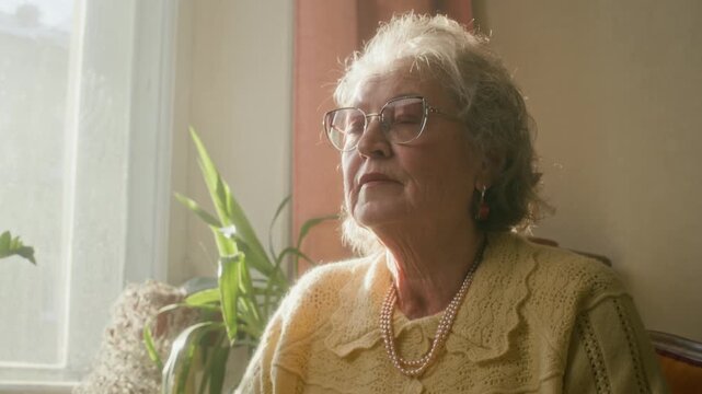 Tilt up shot of thoughtful elderly woman with glasses pausing from reading book recalling past or thinking story over while sitting next to window in retro living room
