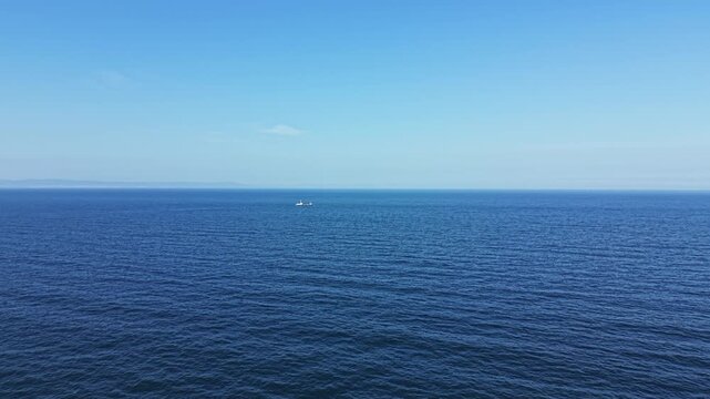 A small boat moves across the open ocean under a clear blue sky. The water appears calm and stretches far into the horizon on a bright day.