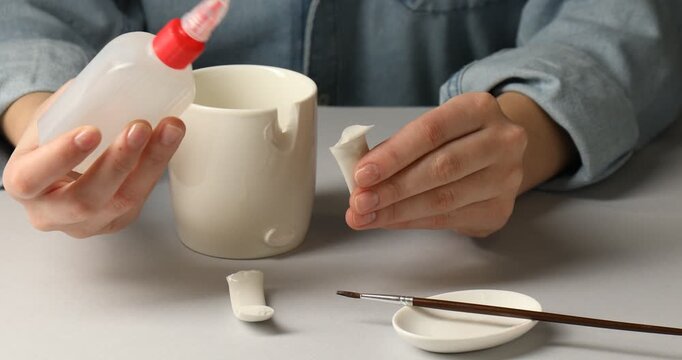 Woman gluing together pieces of broken mug at light grey table, closeup