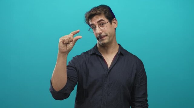 Man pinching fingers to show a tiny amount, wearing glasses and dark shirt against a turquoise studio backdrop with soft lighting; skepticism.
