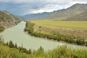 Obraz premium A winding and wide turquoise river crosses a picturesque valley at the foot of the mountain ridge with a summer sunny day. River