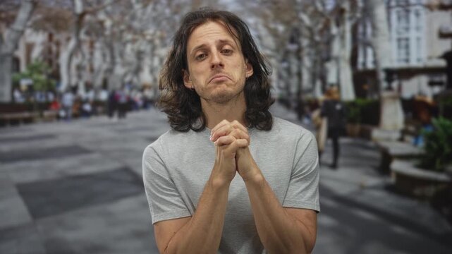 Man with clasped hands and pleading expression on a crowded street with benches and trees, long hair and bare forearms visible; pleading hope.