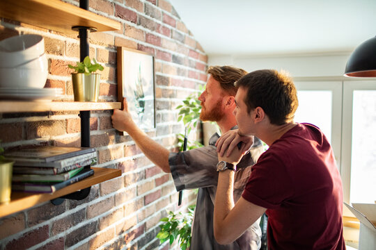 Two men hanging framed art on brick wall at home