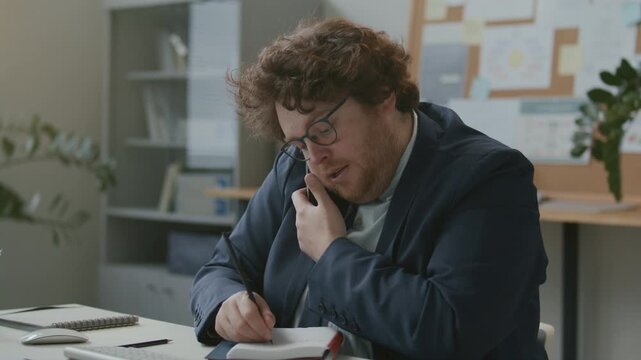 Young male office worker in formal suit and glasses sitting at desk, talking on cell phone and writing notes during busy workday