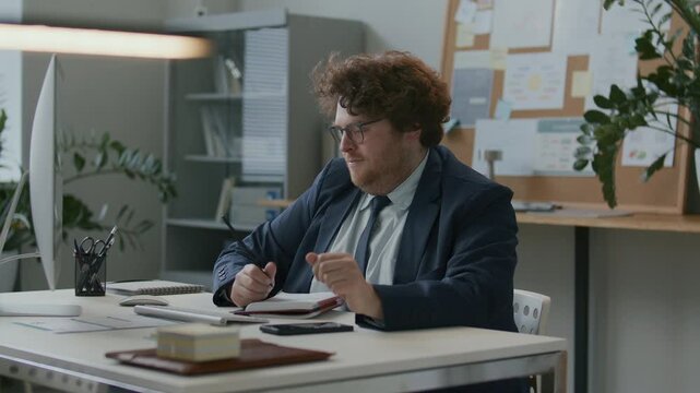 Young businessman using computer and taking notes, then checking message on phone, leaning back in chair and stretching his arms above head, relaxing during break from work