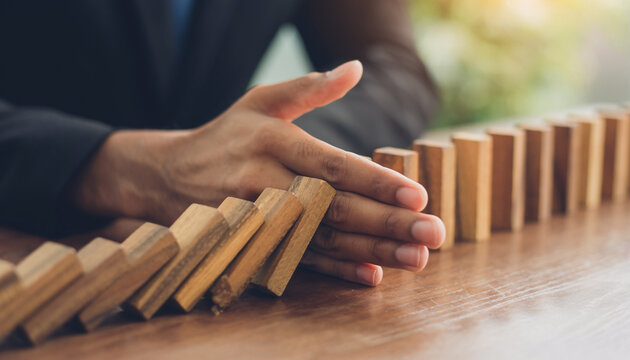 Businessman stopping falling domino blocks with hand, symbolizing risk management, crisis prevention, protection, leadership and strategic control to maintain stability prevent chain reaction failure