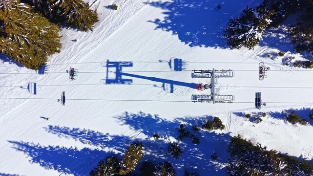 A ski lift is seen from above in a snowy area. The ground is covered in white snow, and there are trees nearby. Shadows fall on the snow, creating patterns on the surface.