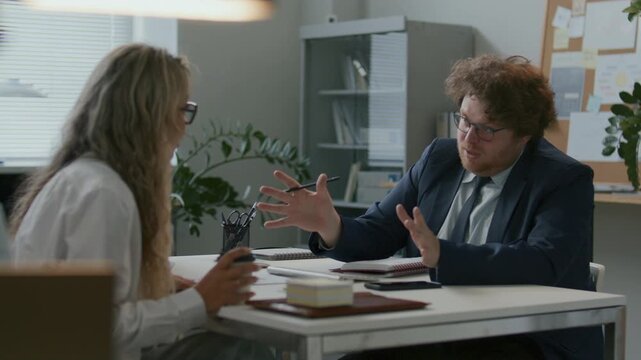 Young businessman sitting at desk across from female colleague, taking notes and discussing work tasks during office meeting