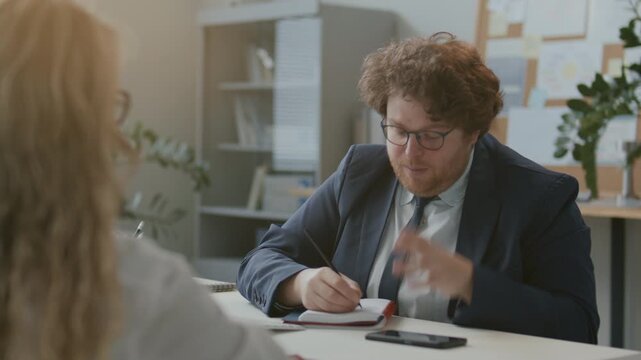 Businessman taking notes and explaining his ideas emotionally during professional meeting with female colleague sitting across desk in sunlit office