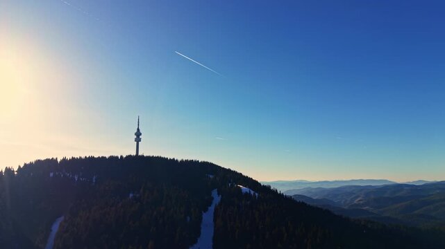 A tall tower is located on a mountain peak as the sun sets in the background. Clear skies and distant hills create a simple scene in nature.