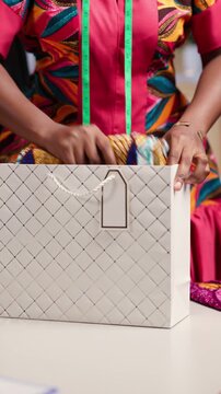 Vertical video, African woman fashion designer entrepreneur packing vibrant ankara print clothes into a shopping bag for her customer