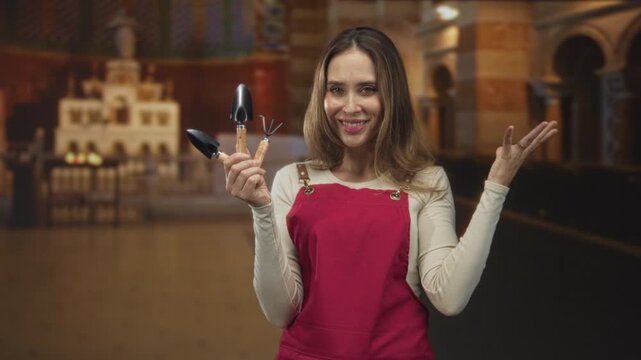 Woman wearing a red apron holding small trowels and a cultivator while smiling and gesturing with her hand inside a building; gardening joy.