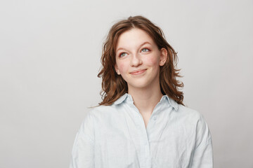 Fototapeta premium Young woman with wavy brown hair wearing white shirt looks up thoughtfully on light background. Concept of contemplation, hope, and casual lifestyle portrait with natural expression.