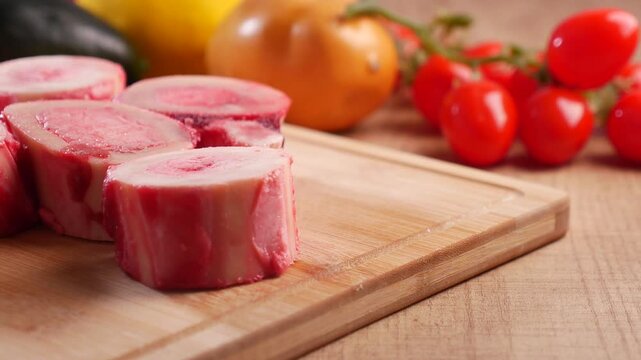 Close-up cinematic video of fresh red raw beef marrow bones on a wooden cutting board, surrounded by vibrant, colorful vegetables, captured with a slow pan.