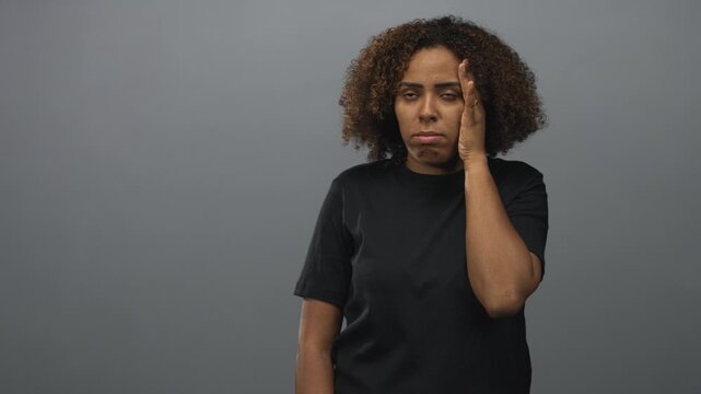 Young woman touching her temple with one hand in a gray studio, eyes rolled and mouth tense, side face visible and curly hair framing cheek; frustration fatigue.
