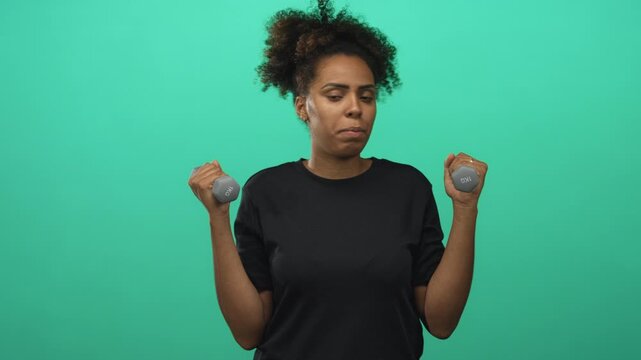 Woman holding small dumbbells and lifting forearms in green studio while wearing black shirt and curly hair pulled up; fitness effort.