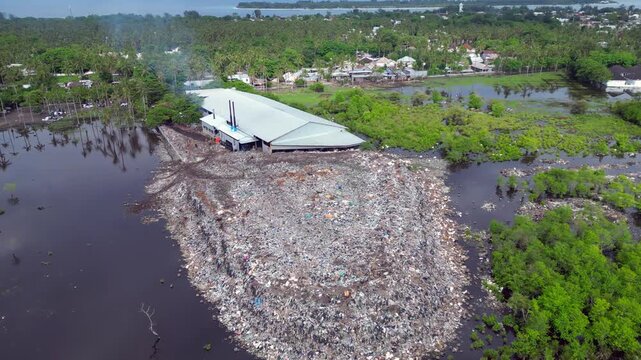 Large rubbish dump spreading into polluted water, showing environmental damage and cows scavenging for food amidst plastic waste. Tremendous aerial view overflight flyover panorama drone