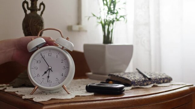 A young woman&rsquo;s hand reaches to turn off a white analog alarm clock on a bedside table, with soft natural light, a plant, and personal items in the background, close-up shot.