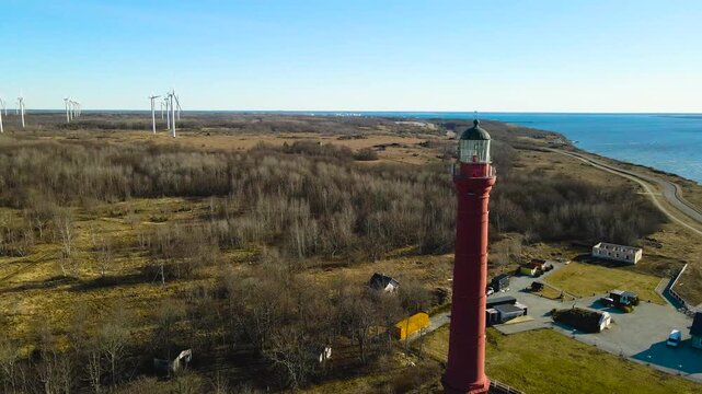 Beautiful aerial footage flying around the iconic red pakri lighthouse on the coastline, revealing a modern wind farm generating renewable energy on a sunny day in paldiski, estonia