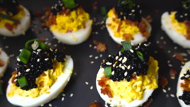 Delicious deviled eggs topped with black caviar, crispy bacon bits, sesame seeds, and fresh green onions served on a dark platter, creating an elegant and savory party appetizer