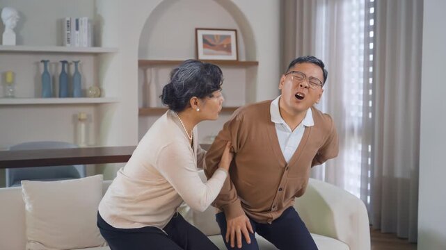 Fatigue senior man grandpa sitting at sofa couch, feel lower back pain, hurt sudden back ache, mature female wife help him standing, osteoporosis, arthritis, lumbar spine issues aging health condition