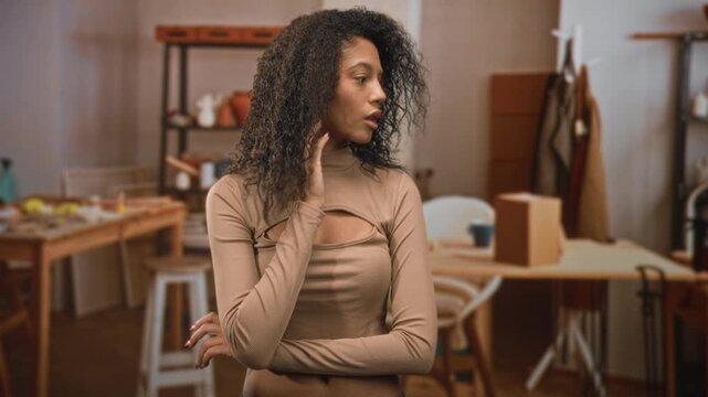 Teenage woman with curly hair touches her neck and crosses arms while standing near a worktable with boxes and ceramic vases in a studio; contemplation.