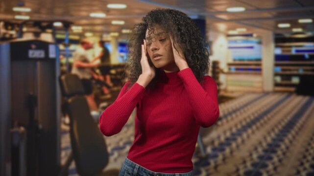 Woman in red sweater pressing temples with both hands beside a gym weight machine on patterned carpet inside a building; stress fatigue.