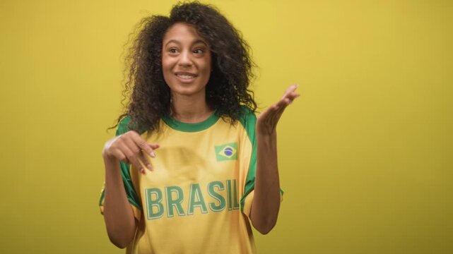 Teenage woman with curly hair wearing brasil football t shirt with brazil flag patch claps hands and smiles broadly in studio; joy pride.