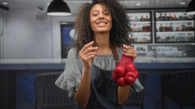 Woman with curly hair wearing an apron holding a net bag of red tomatoes while pinching fingers toward camera at a building bar counter; cheerful hospitality.