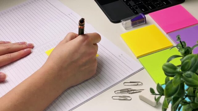 A close-up of a woman's hands writing a note on a colored sticky note at a desk next to a notebook, laptop, and stationery. Conceptually, this represents planning, task organization, and productivity.