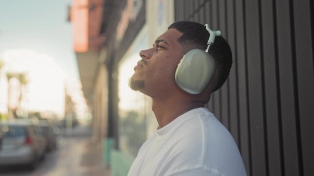 Hispanic man with beard wearing large over ear headphones stands with eyes closed, head tilted back against a storefront wall on a street; calm reflection.