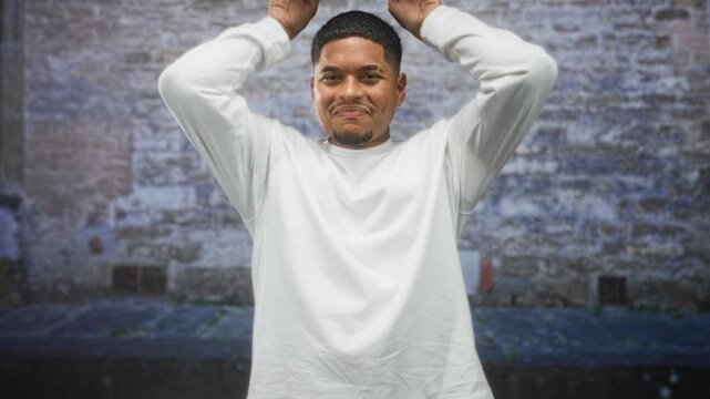 Hispanic man with beard, hands on head gesture and slight smile framed against a weathered brick wall on a street; playful confidence.