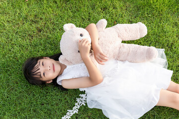 A little girl in a white dress holding a stuffed bear while lying on the grass © mei