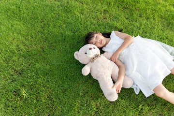 A little girl in a white dress holding a stuffed bear while lying on the grass © mei