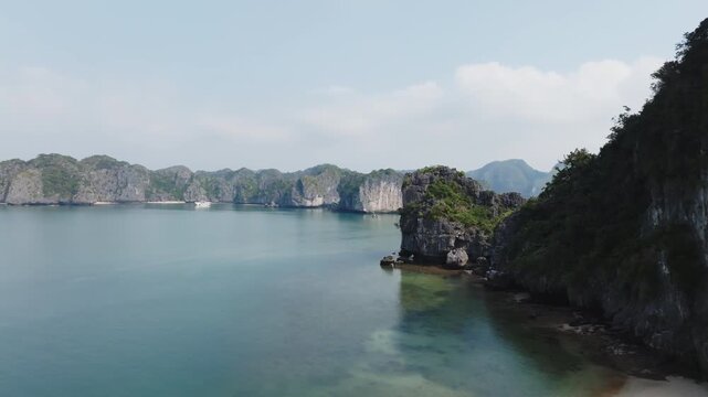 Cat Ba Vietnam drone view of a rocky islet beside calm open water, with distant limestone walls and hazy hills.