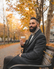 Fototapeta premium Autumnal Contemplation: A man, wrapped in a coat, pauses on a park bench, cradling a warm beverage while the crisp autumn air surrounds him. Golden leaves of the trees cast a warm glow.