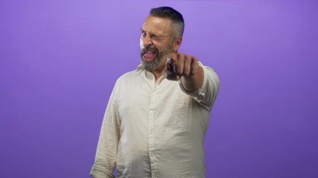 Middle aged hispanic man with grey beard and rolled sleeves points finger to camera in purple studio; playful invitation to engage.
