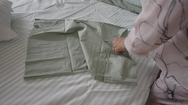 Woman hands putting light green cotton pillowcase on white pillow on striped bedsheet in bedroom.